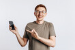 © Daria Lukoiko - Young handsome man holding a smartphone and smiling. The guy cheerfully points his finger at the phone. Student, worker posing for photo isolated on gray background. Place for advertising.
