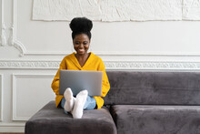 Smiling African American millennial woman with afro hairstyle wear yellow cardigan sitting on sofa, resting, looking at camera webcam and talking on a video call or skype with friends, watching movie