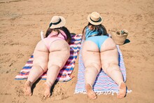 Backview Of Two Plus Size Overweight Sisters Twins Women Relaxing Lying On A Towel At The Beach On Summer Holidays