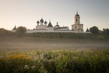 A Russian Orthodox monastery on misty sunrise. A meadow with yellow flowers in the foreground. Clear colourful sky. No people