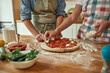 For cheesy taste. Cropped shot of couple making pizza together at home. Man in apron adding, applying tomato sauce on the dough while woman adding mozzarella cheese. Hobby, lifestyle