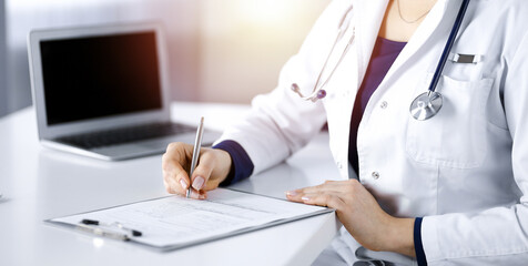 Unknown woman-doctor is prescribing some medication for her patient, using a clipboard, while sitting at the desk in her sunny cabinet. Female physician with a stethoscope, close up. Perfect medical