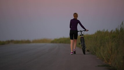 Wall Mural - Young woman rolls her bike near herself on the countryside road at twilight, back view