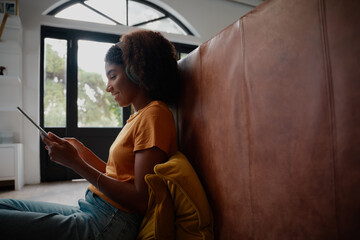 Wall Mural - Side view of young african woman sitting on floor using digital tablet with headphones while on video call