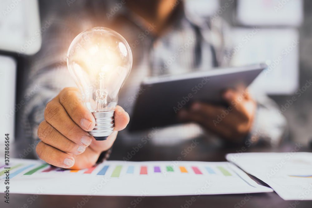 Successful business idea and creative innovation concept, close up business man holding lighting bulb and tablet with financial graph on table - obrazy, fototapety, plakaty 