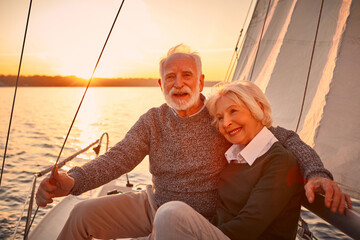 Portrait of a beautiful and happy senior couple in love hugging and enjoying amazing sunset while sitting on the side of sailboat or yacht deck floating in sea