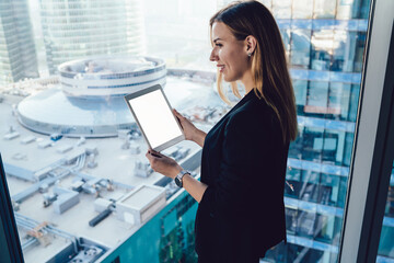 Wall Mural - Back view of successful female entrepreneur in formal clothing smiling near panoramic window holding digital tablet with copy space area for business website, happy corporate boss with blank touch pad