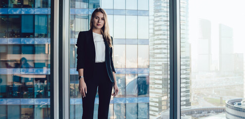 Wall Mural - Half length portrait of pretty blonde business woman in trendy elegant formal wear standing near window in office interior, confident beautiful 30s female executive manager in suit looking at camera