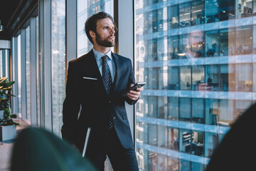 Wall Mural - Serious caucasian male entrepreneur in formal wear looking outside from panoramic window in office sending message on mobile phone, confident businessman holding smartphone using app for communication