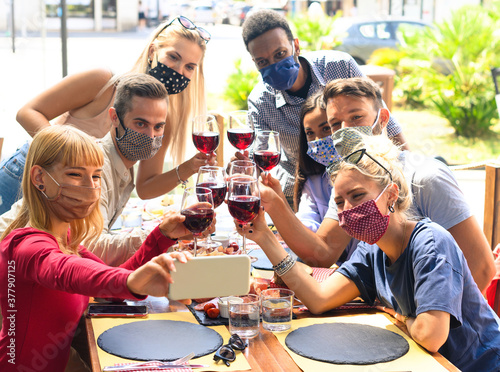 Friends with face mask taking selfie while toasting red wine at restaurant