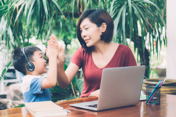 Wall Mural - Young asian mother and son using laptop computer for study and learning and hight five together at home, boy wearing headphone for e-learning with distancing, mom support child, education concept.
