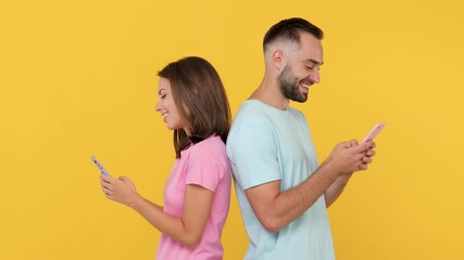 Wall Mural - Young couple friends bearded man woman 20s in basic t-shirt standing back to back isolated on yellow background studio. People emotion lifestyle concept Look at gadget using mobile cell phone chatting