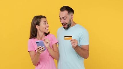 Wall Mural - Young couple friends woman man in basic t-shirts posing isolated on yellow background studio. Using mobile phone hold credit bank card online shopping fun expressive doing winner gesticulating hands