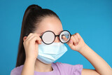 Fototapeta Desenie - Little girl wiping foggy glasses caused by wearing medical face mask on blue background. Protective measure during coronavirus pandemic