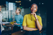Pensive African American Woman Holding Optical Glasses In Hand And Thoughtful Thinking On Information, Contemplative Female With Dark Skin Standing In Coworking Space And Pondering Looking Away