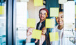 © BullRun - Cheerful male and female colleagues analyzing information from memo sticks making notes of interesting ideas and laughing, happy diverse colleagues enjoying together brainstorming near glass board
