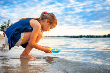 Side view of little lovely caucasian girl playing with tiny rubber yellow ducks in small blue pool, standing on beach sand. Attractive kid having fun, enjoying holidays at sea, summertime.