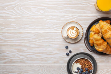 Traditional morning breakfast food on rustic white wooden background. Fresh croissants, granola with yoghurt and berries, coffee cappuccino and orange juice. Breakfast meal setting, space for text.