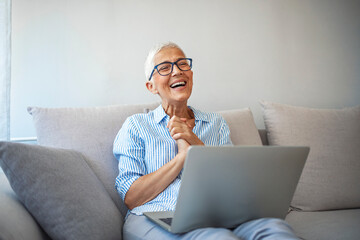 Senior woman making video call on laptop , waving at screen, chatting online with people who distance. Cropped shot of a happy senior woman waving hello while on a video call on her couch at home