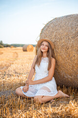 Poster - Attractive barefoot girl in white dress and a straw hat near the bale of straw in the field after the bread harvest