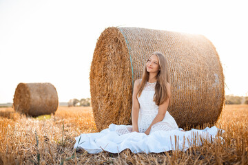 Canvas Print - Rural scene of two bales of straw with a young girl on the foreground in white dress sitting under the bale and looking aside