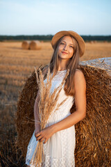 Sticker - Young blond girl leaning on the bale of straw and holding a bouquet of dried flowers catching last warm sun rays of a setting sun