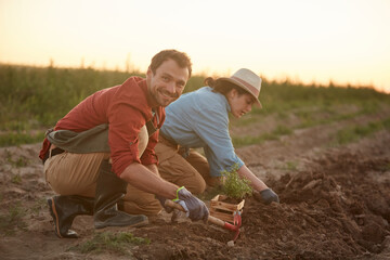 Full length portrait of two people working in field at vegetable plantation, focus on young man planting saplings in foreground and smiling at camera, copy space