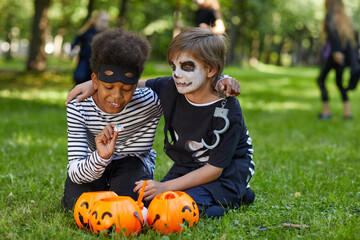 Wall Mural - Full length portrait of two boys wearing Halloween costumes sitting on green grass outdoors with candy buckets, copy space