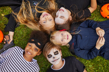 Wall Mural - Above view at multi-ethnic group of kids wearing Halloween costumes while lying on grass and looking at camera