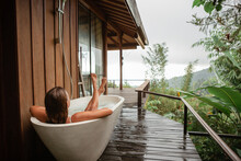 Back View Woman Pampering Her Body In Water While Lie In Bath Tube Outdoor With Jungle And Mountains View