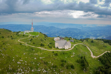 Drone panorama of Top of Urslja gora or Plesivec in the Koroska region of slovenia on a summer day. Visible grass, church of st. Ursula, and TV antenna on the top, with Pohorje mountains in the backgr