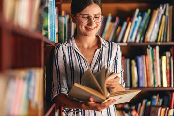 Young smiling attractive college girl leaning on book shelves in library and searching for material for homework.