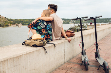 Wall Mural - Young couple with electric scooter on vacation having fun in the city, while sitting by the river.