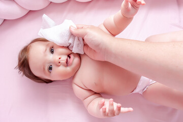 hygiene - young mom wiping the baby skin body and face with wet wipes carefully on pink background. 