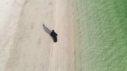 Wall Mural - Muslim woman walking by the seaside wearing hijab abaya dress aerial view