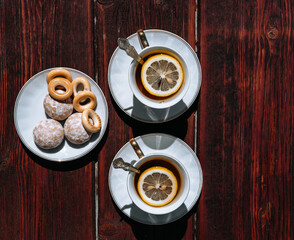 Beautiful white ceramic tableware for tea drinking. Two white cups of black tea with lemon on a wooden table, and delicious bagels and gingerbread in a saucer.