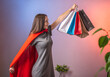 © Tatiana - A young determined woman in a superhero cape and holding up shopping bags. Concept of sales and shopping