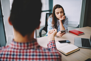 Wall Mural - bored female employee in smart casual outfit sitting at office desktop with blurred male colleague, 
