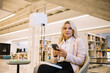© BullRun - Caucasian smart casual woman holding cellphone technology and looking at camera, portrait of confident female student with modern smartphone device posing near table with education literature