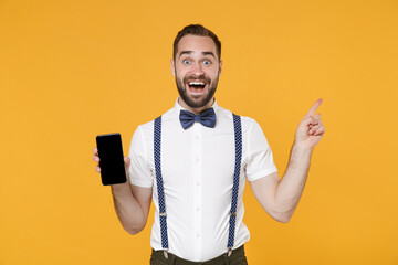 Sticker - Excited young bearded man 20s in white shirt bow-tie suspender pointing index finger up hold mobile cell phone with blank empty screen mock up copy space isolated on yellow background studio portrait.