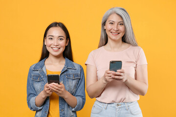 Canvas Print - Smiling funny family asian female women gray-haired mother brunette daughter in casual clothes posing using mobile cell phone typing sms message isolated on yellow color background studio portrait.