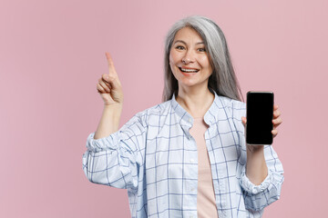 Canvas Print - Funny gray-haired asian woman in white checkered shirt pointing index finger up hold mobile phone with blank empty screen mock up copy space isolated on pastel pink colour background studio portrait.