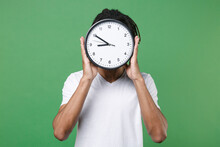 Young African American Man Guy With Dreadlocks 20s Wearing White Casual T-shirt Posing Holding In Hands Covering Face With Round Clock Isolated On Green Color Wall Background Studio Portrait.