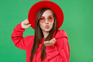 Wall Mural - Pretty charming young brunette woman 20s in bright red casual streetwear hoodie eyeglasses hat posing blowing sending air kiss looking camera isolated on green color wall background studio portrait.