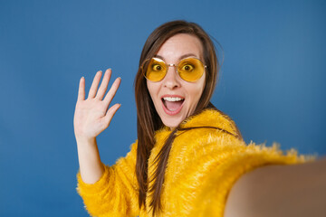 Wall Mural - Close up of excited young brunette woman 20s in yellow fur sweater eyeglasses posing doing selfie shot on mobile phone waving greeting with hand isolated on blue color wall background studio portrait.