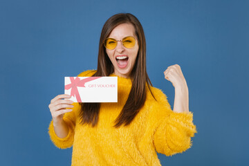 Wall Mural - Happy screaming young brunette woman 20s wearing yellow fur sweater eyeglasses posing holding gift certificate doing winner gesture looking camera isolated on blue color background studio portrait.