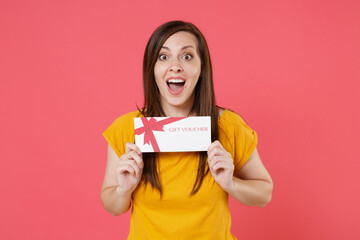 Wall Mural - Excited surprised young brunette woman 20s in yellow casual t-shirt posing holding in hands gift certificate keeping mouth open looking camera isolated on pink color wall background studio portrait.