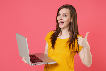 Wall Mural - Funny cheerful young brunette woman 20s wearing yellow casual t-shirt posing and working on laptop pc computer showing thumb up looking camera isolated on pink color wall background studio portrait.