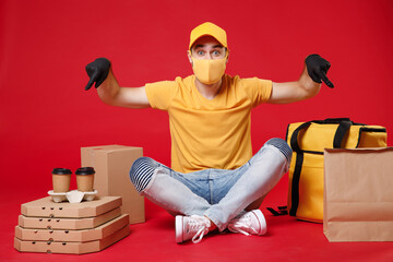 Wall Mural - Delivery employee man in yellow cap face mask gloves tshirt sit at thermal takeaway food bag backpack pizza boxes work courier service during quarantine coronavirus covid-19 isolated on red background