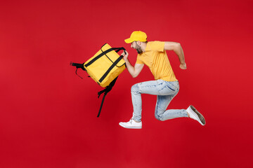 Wall Mural - Full length delivery employee man guy male 20s in yellow cap t-shirt uniform thermal food bag backpack work courier service during quarantine covid-19 virus jumping isolated on red background studio.